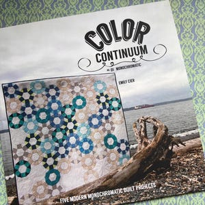 May include: A book cover titled "COLOR CONTINUUM" featuring a quilt with blue, teal, and beige geometric patterns. The cover also displays a photo of a beach with driftwood and the text "FIVE MODERN MONOCHROMATIC QUILT PROJECTS."