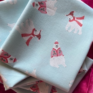 Puede incluir: Una tela de color azul claro con un patrón repetitivo de osos polares blancos. Los osos llevan gorros de invierno rojos y blancos, bufandas y chaquetas rosas adornadas con hojas de acebo verdes. La tela tiene una textura tejida visible.