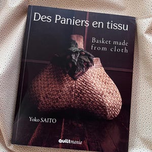 May include: A book titled "Des Paniers en tissu" with the subtitle "Basket made from cloth" by Yoko SAITO. The cover features a woven basket with a wooden form and green foliage. The background is a light beige fabric with small, evenly spaced dots.