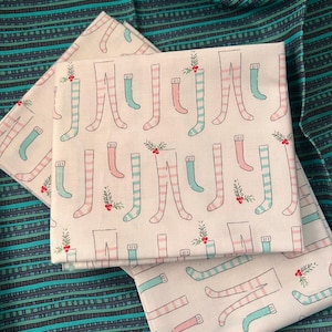 May include: A folded piece of off-white fabric featuring a repeating pattern of pink and teal striped stockings and small sprigs of holly with red berries. The fabric is layered over a dark teal and black striped textile with a textured appearance.