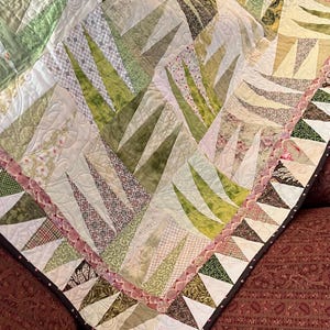 May include: A handmade quilt with a patchwork design featuring various shades of green, cream, and brown. The quilt has a repeating pattern of triangular shapes and is bordered with a pink ruffle and a dark brown binding. The quilt is on a red patterned surface.
