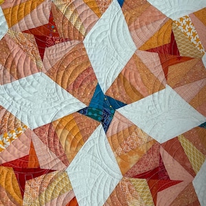 Op de afbeelding: Close-up van een kleurrijke quilt met een sterrenpatroon. De quilt heeft een mix van oranje, perzik, rood, wit en blauwgroene stoffen. Het ontwerp bevat gebogen lijnen en geometrische vormen, wat een textuur en visueel aantrekkelijk textiel creëert.