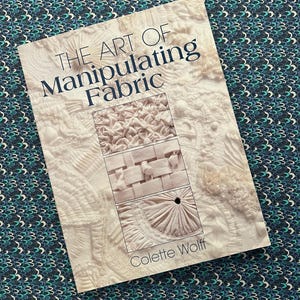 May include: A book titled "THE ART OF Manipulating Fabric" by Colette Wolff. The cover features close-up images of fabric manipulation techniques. The background is a patterned fabric with teal, blue, and white colors.