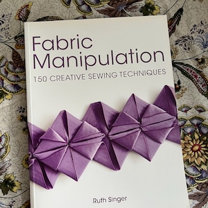 May include: A book titled "Fabric Manipulation" with the subtitle "150 Creative Sewing Techniques" by Ruth Singer. The cover features a white background with a purple ribbon design. The book is on a floral patterned surface.