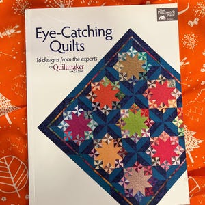 Puede incluir: Un libro titulado "Eye-Catching Quilts" con 16 diseños de Quiltmaker Magazine. La portada muestra un diseño de edredón con patrones de estrellas coloridas sobre un fondo azul oscuro. El libro está sobre una tela naranja con patrones de árboles y hojas blancas.