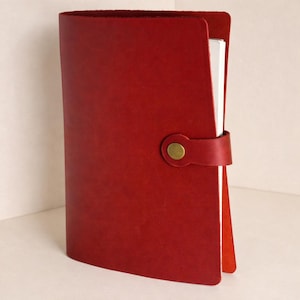 May include: A red leather journal with a curved spine and a strap closure secured by a brass button. The journal's pages are visible on the right side. The leather has a smooth texture. The journal is a classic stationery item.