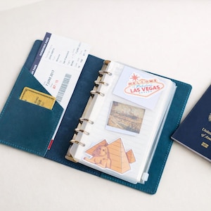 May include: Open teal leather travel journal with a boarding pass, credit card, and stickers. The stickers include a Las Vegas sign, a photo, and a pyramid. A passport is visible on the right.