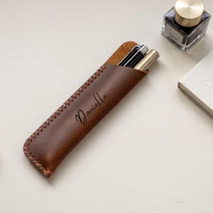 May include: A dark brown leather pen sleeve featuring visible whipstitching along the edge and the name Danielle embossed in black script. The sleeve contains two pens, one black and one gold, and is positioned near a glass ink bottle.