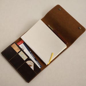 以下が含まれることがあります： クリーム色のノートパッド、ペン、ポケットが付いた、開いた茶色の革製ジャーナル。ジャーナルは開いており、空白のノートパッド、濃い茶色のペン、カードや搭乗券が入ったいくつかのポケットが表示されています。ジャーナルは2つのスナップ留めで閉じられています。