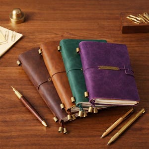 Peut inclure: Plusieurs carnets en cuir de couleurs marron, vert et violet, avec des détails en laiton. Les carnets sont posés sur une surface en bois, accompagnés de stylos. Les carnets mesurent environ 15 cm de haut.