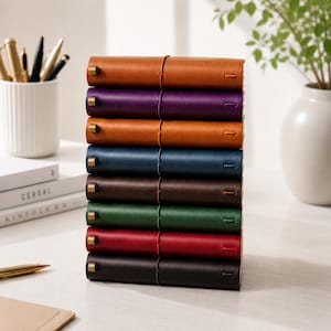Op de afbeelding: Een stapel kleurrijke leren journals in verschillende tinten, waaronder bruin, paars, blauw, groen, rood en zwart. Elk journal is gebonden met een leren band en een messing sluiting. De journals zijn netjes op een wit oppervlak gestapeld.
