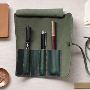 Pode incluir: Um estojo de couro verde-oliva com quatro compartimentos verticais, três dos quais contêm canetas. O estojo inclui uma tira longa de couro para fechar. Perto encontram-se um caderno castanho, um prato de cobre com clipes e papel.