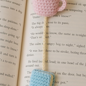 May include: A handmade crochet bookmark featuring a pink coffee cup with brown 'coffee' and a light blue book. The bookmark is placed on the pages of a book, adding a touch of whimsy to reading.