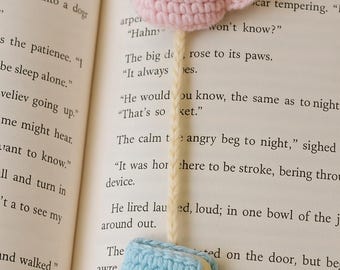 Puede incluir: Un marcapáginas de ganchillo hecho a mano con una taza de café rosa con 'café' marrón y un libro azul claro. El marcapáginas se coloca en las páginas de un libro, añadiendo un toque de fantasía a la lectura.