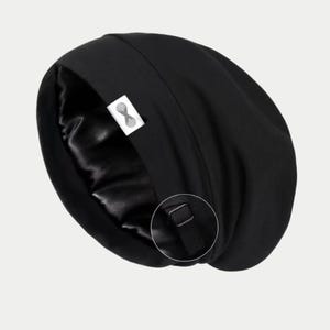 May include: Black satin-lined sleep cap. The exterior is a solid black fabric, while the interior is a shiny black satin. A small white tag with a logo is visible. The cap has an adjustable strap.