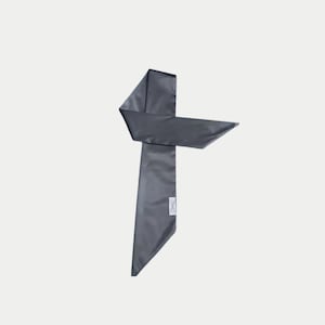 May include: A dark gray satin scarf, folded and tied in a unique shape. The scarf has a rectangular label at the bottom. The scarf is a solid color and appears to be made of a smooth, shiny material.