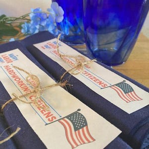 Könnte beinhalten: Eine patriotische Tischdekoration zum Tag der Arbeit mit zwei kobaltblauen Trinkgläsern, marineblauen Servietten und Etiketten mit amerikanischen Flaggen. Auf den Etiketten steht "Happy Labor Day! Hardworking Citizens."