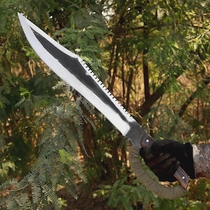 May include: A large silver machete with a black blade edge and serrated spine. The handle is wrapped in green fabric and has a dark wood grip. The machete is set against a backdrop of green foliage.