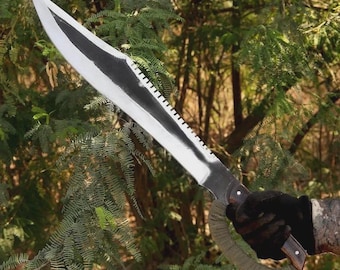 May include: A large, silver machete with a black blade edge and serrated spine. The handle is wrapped in green fabric and has a dark wood grip. The machete is held against a backdrop of green foliage.