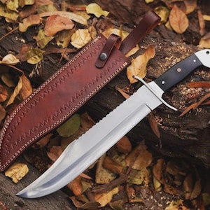 Puede incluir: Un cuchillo grande de acero inoxidable con borde dentado y mango negro, sobre un lecho de hojas y madera. El cuchillo tiene una funda de cuero marrón con costuras decorativas y un lazo para cinturón.
