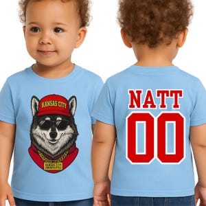A young child wearing a blue t-shirt with a graphic of a wolf wearing sunglasses and a red bandana, along with the text "KANSAS CITY" and "COMBOSTREET" printed on it.