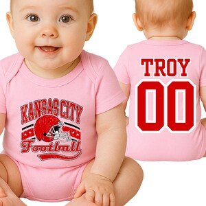 A baby wearing a pink onesie with a Kansas City football jersey and the name "Troy" printed on it.