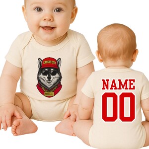 A baby wearing a white onesie with a graphic of a wolf wearing a red hat and a red bandana, sitting on the floor.