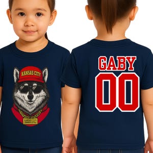 A young girl wearing a navy blue t-shirt with a graphic of a wolf wearing sunglasses and a red hat, along with the text "KANSAS CITY" and "KANSAS CITY COWBOYS".