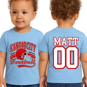 A young child wearing a light blue t-shirt with a graphic of a football helmet and the text "KANSAS CITY FOOTBALL" printed on it. The child's name, "MATT 00", is also displayed on the back of the shirt.