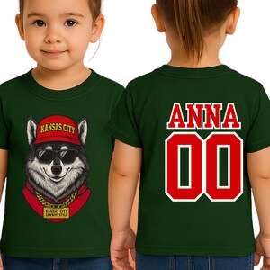 A green t-shirt with a graphic of a wolf wearing sunglasses and a red hat, along with the text "KANSAS CITY" and "KANSAS CITY COMBBOSTYLE". The back of the shirt has the name "ANNA" and the number "00" printed on it.