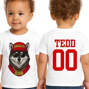 A white t-shirt with a graphic of a wolf wearing sunglasses and a red bandana, along with the text "KANSAS CITY" and "KANSAS CITY WORKS STYLE". The back of the t-shirt has the name "TEDD" and the number "00" printed on it.