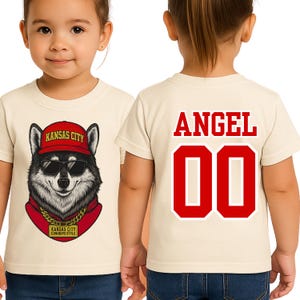 A young girl wearing a white t-shirt with a graphic of a husky dog wearing sunglasses and a red hat, along with the text "KANSAS CITY" and "KANSAS CITY COWBOYS STYLE".
