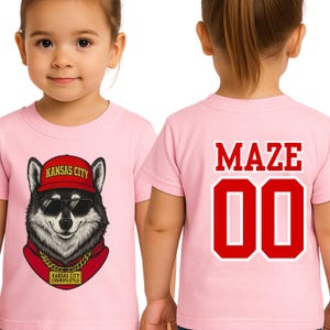 A young girl wearing a pink t-shirt with a graphic of a wolf wearing sunglasses and a red hat, along with the text "KANSAS CITY" and "KANSAS CITY COWBOYS".