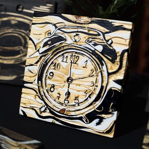 May include: Square canvas art depicting a clock face. The design uses gold, black, and white paint with a textured finish. The clock hands indicate around 11 o'clock. The artwork has a vintage aesthetic. The measurements are in centimeters.