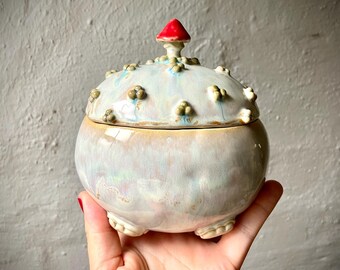 Puede incluir: Un tarro de cerámica redondo con tapa y acabado brillante e iridiscente en tonos azul pálido, rosa y crema. La tapa presenta un pequeño pomo rojo en forma de seta y varias protuberancias texturizadas en relieve sobre su superficie.