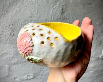 Puede incluir: Un pequeño cuenco de cerámica sostenido en la mano con un interior amarillo brillante y un exterior texturizado de color blanco roto. El cuenco presenta una serie de pequeños recortes circulares a lo largo del borde superior y una flor rosa esculpida en relieve con hojas verdes.