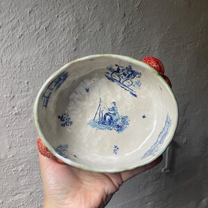 Könnte beinhalten: Eine Keramikschale mit cremefarbener Innenseite, verziert mit blauen Illustrationen im Toile-Stil, die eine Person an einer Staffelei und eine Person zu Pferd zeigen. Der äußere Rand ist mit dreidimensionalen roten Erdbeeren verziert.