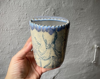 Puede incluir: Una maceta de cerámica para pared con base color crema y una ilustración de pulpo azul. El borde superior presenta una textura de cuentas decorativa con un efecto de goteo de esmalte azul. El recipiente tiene una parte trasera plana.