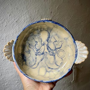 Könnte beinhalten: Eine runde Keramikschale mit zwei muschelförmigen Seitengriffen, die im cremefarbenen Inneren eine blaue Tintenfischillustration mit Gabel, Löffel und Teetasse zeigt. Die Schale hat einen blau bemalten Rand und ein gesprenkeltes Finish.