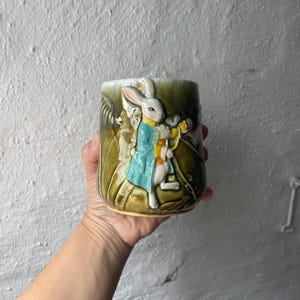 Könnte beinhalten: Ein Keramikbecher mit glänzender olivgrüner Glasur und einem erhabenen, dreidimensionalen Relief eines weißen Kaninchens, das einen blauen Mantel und eine gelbe Weste trägt. Das Kaninchen ist in einer Waldumgebung mit texturiertem Laub dargestellt.