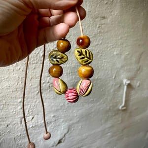Puede incluir: Un collar artesanal con cuentas de cer&aacute;mica en tonos marr&oacute;n, amarillo y rosa. Las cuentas, con forma de hoja y redondas, est&aacute;n ensartadas en un fino cord&oacute;n marr&oacute;n claro. El collar est&aacute; sostenido por una mano. Aproximadamente 40 cm de largo.