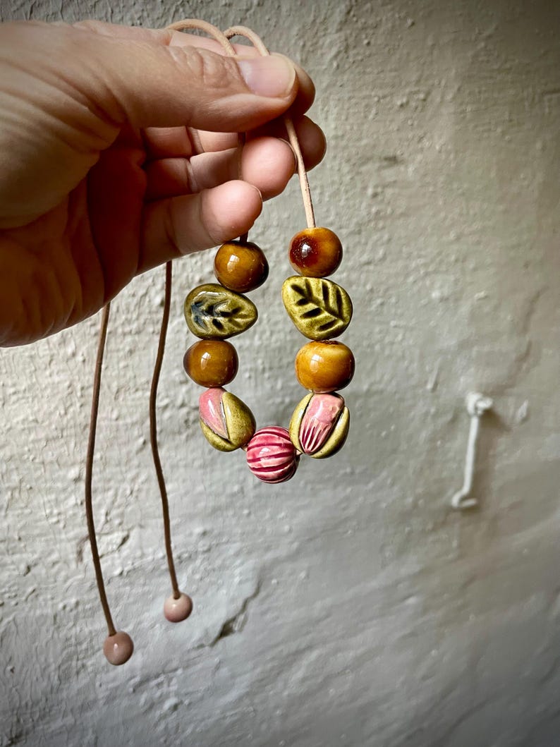 Puede incluir: Un collar artesanal con cuentas de cer&aacute;mica en tonos marr&oacute;n, amarillo y rosa. Las cuentas, con forma de hoja y redondas, est&aacute;n ensartadas en un fino cord&oacute;n marr&oacute;n claro. El collar est&aacute; sostenido por una mano. Aproximadamente 40 cm de largo.