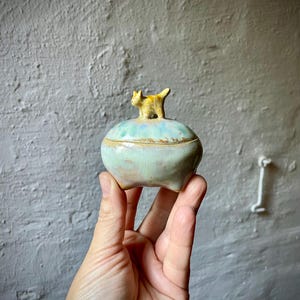 Könnte beinhalten: Kleine, handgefertigte Keramikdose mit Deckel. Die Dose ist hellgrün mit einem blau-grünen Rand und einer kleinen, gelben Katzenfigur oben. Die Dose wird in der Hand einer Person gehalten.