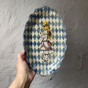 Könnte beinhalten: Eine ovale Keramik-Servierschale mit gewelltem Rand und einem blau-cremefarbenen Rautenmuster. In der Mitte befindet sich eine detaillierte Illustration einer Figur mit gelber Krone und Zepter, bekleidet mit einem weißen, gerüschten Outfit.