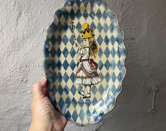 Puede incluir: Una fuente de cerámica ovalada con borde festoneado, que presenta un patrón de rombos en azul y crema. El centro muestra una ilustración detallada de una figura con corona amarilla y cetro, vestida con un atuendo blanco con volantes.