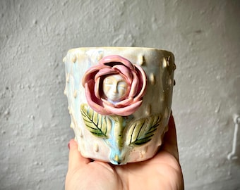 Puede incluir: Una taza de cerámica con un acabado brillante e iridiscente y puntos texturizados en relieve. Una rosa rosa esculpida en tres dimensiones está fijada en la parte frontal, con un pequeño rostro humano sereno en el centro de los pétalos.