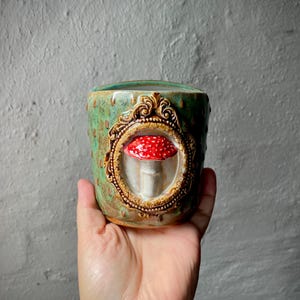 Könnte beinhalten: Ein Keramikbecher mit gesprenkelter grüner und brauner Glasur, der einen erhabenen, dreidimensionalen rot-weißen Pilz zeigt, welcher in einem verzierten, goldfarbenen dekorativen Rahmen auf der Außenfläche des Gefäßes zentriert ist.