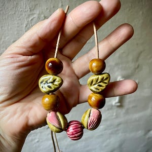 Puede incluir: Un collar hecho a mano con cuentas de cer&aacute;mica en tonos marr&oacute;n, rosa y verde. Las cuentas incluyen dise&ntilde;os redondos, en forma de hoja y a rayas, ensartadas en un cord&oacute;n marr&oacute;n claro. El collar est&aacute; sostenido por una mano.