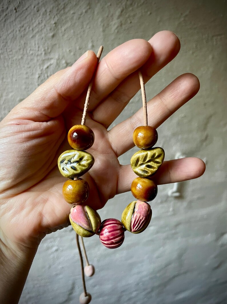 Puede incluir: Un collar hecho a mano con cuentas de cer&aacute;mica en tonos marr&oacute;n, rosa y verde. Las cuentas incluyen dise&ntilde;os redondos, en forma de hoja y a rayas, ensartadas en un cord&oacute;n marr&oacute;n claro. El collar est&aacute; sostenido por una mano.