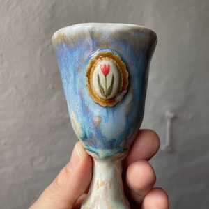 Könnte beinhalten: Ein handgefertigter Keramikbecher mit blau-weißer Glasur. Der Becher hat ein kleines, rundes Medaillon mit einem roten Tulpenmuster. Der Sockel ist braun und der Stiel ist weiß und beige. Der Becher ist etwa 10 cm hoch.
